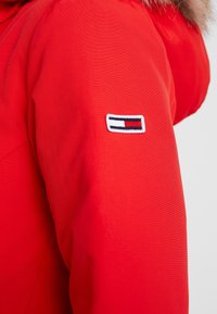 Rood bovenkledingstuk met een gladde textuur en een capuchon met bontkraag; bevat een wit patch met blauwe en rode strepen op de arm.