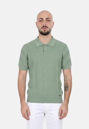 Uomo con barba e tatuaggi che indossa una polo a maniche corte verde chiaro con trama e pantaloni bianchi, in piedi davanti a uno sfondo semplice.