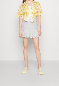 Chemise à manches courtes à fleurs jaunes nouée à l'avant, débardeur blanc, jupe lavande brodée avec une bordure à volants, et des chaussures à enfiler perforées jaunes.