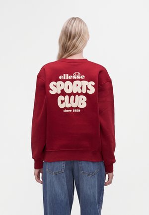 Човек с дълга руса коса, носещ червен суитшърт с бял текст ""ellesse SPORTS CLUB since 1959"" на гърба и сини дънки.