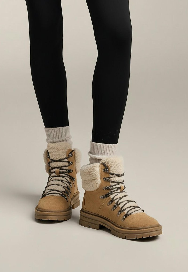 Tan suede ankle boots with white cozy fur collar. Thick black laces and metal eyelets, textured rubber sole, worn with black leggings.
