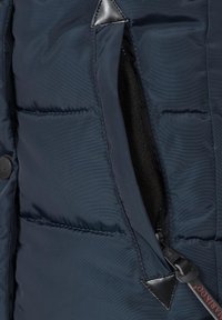 Námorně modrá péřová bunda s kapsou na zip, s měkkou černou podšívkou a trojúhelníkovým koženým akcentem blízko otvoru.