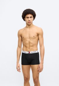 Μαύρα boxer briefs με λευκή ελαστική μέση που φέρει το λογότυπο "TOMMY HILFIGER". Το μοντέλο έχει κοντά κυματιστά μαλλιά και ουδέτερη έκφραση.