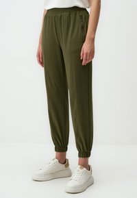 Olijf groene elastische taillebroek met taps toelopende boorden, zijrits zak en glad, lichtgewicht stof. Draagt met witte sneakers.