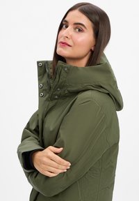 Olijfgroene jas met een hoge kraag en een capuchon, voorzien van drukknoopsluitingen en elastische manchetten. Gladde textuur met een aansluitende pasvorm.