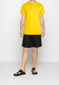Žluté polo tričko s límečkem, černé šortky a černé sandály bez podpatku. Jednoduchý design s krátkými rukávy a bez viditelných vzorů.