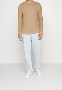 Suéter marrón claro con textura acanalada, jeans azul claro ajustados y zapatillas blancas, posando sobre un fondo liso.