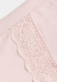 Růžová tkanina s texturovaným krajkovým lemem, který má květinové vzory a vlnité okraje. Hladký povrch, jemný lesk, delikátní detaily.