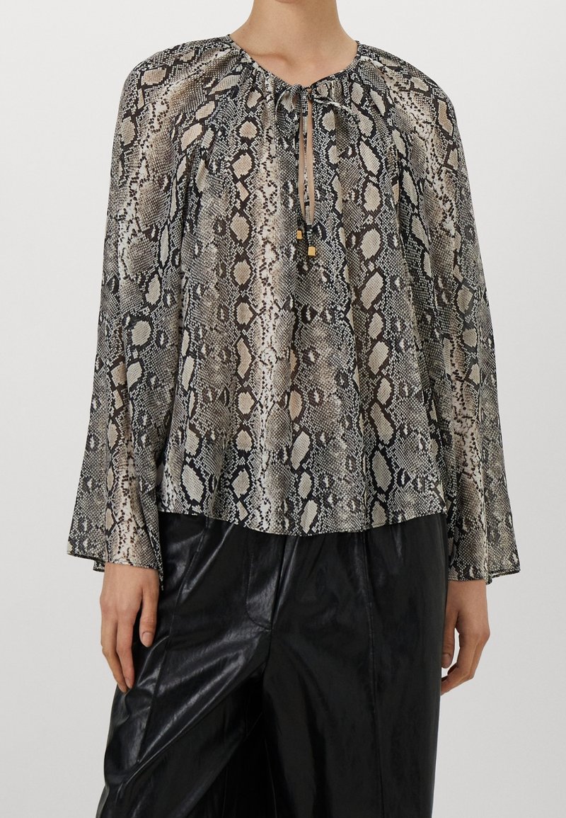 Blouse ample à manches longues avec imprimé serpent et col à nouer, associée à un pantalon en cuir noir, portée par une personne debout sur un fond uni.