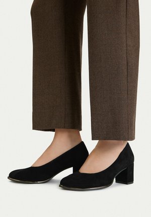 Nohy v černých semišových botách s blokovým podpatkem, doplněné hnědými kostkovanými kotníkovými kalhotami na neutrálním pozadí.