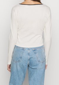 Dlouhý rukáv bílý žebrovaný top s černým výstřihem, v kombinaci s vysokými lehce modrými denimovými kalhotami s volným střihem a zadními kapsami.