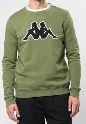 Mand iført olivengrøn sweatshirt med sort silhuetlogo af to personer, der sidder ryg mod ryg på brystet, kombineret med sorte bukser.