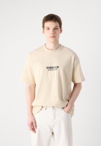Jasno beżowa bawełniana koszulka z krótkim rękawem, z czarnym logo "BARBOUR INTERNATIONAL" z przodu. Prosty, luźny krój.