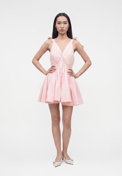 Γυναίκα στέκεται με τα χέρια στη λεκάνη φοράει αμάνικο ανοιχτό ροζ φόρεμα με δεμένες τιράντες στους ώμους και μπεζ παπούτσια με χαμηλό τακούνι.