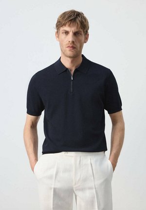 Hombre con cabello castaño claro vistiendo una camiseta polo negra de manga corta y pantalones plisados blancos, de pie con las manos en los bolsillos frente a un fondo liso.