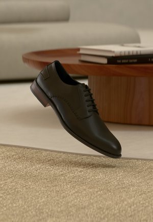 Chaussure Oxford en cuir noir avec des détails perforés flottant au-dessus d'un tapis beige texturé, une table ronde en bois et une pile de livres en arrière-plan.