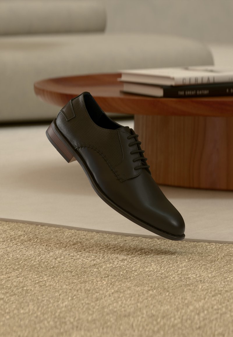 Chaussure Oxford en cuir noir avec des détails perforés flottant au-dessus d'un tapis beige texturé, une table ronde en bois et une pile de livres en arrière-plan.