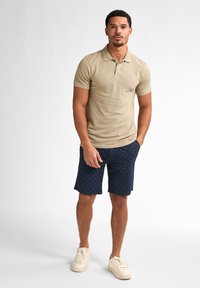 Beige polo tričko s krátkým rukávem a límečkem, spárované s námořnickými kraťasy s texturovaným vzorem a bílými teniskami.