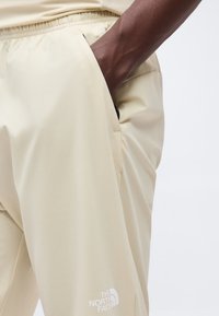 Πρόσωπο με σκούρο δέρμα με το χέρι στην τσέπη, φορώντας μπεζ παντελόνι με ελαστική μέση που φέρει το λογότυπο The North Face στο αριστερό μηρό.