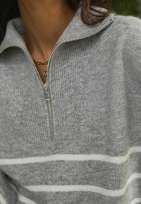 Pull en gris à zip fabriqué dans un matériau doux, avec une texture côtelée et des rayures blanches horizontales sur le corps. Détail de fermeture éclair argenté visible.