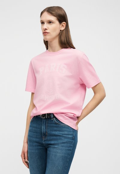 Νεαρή γυναίκα που φοράει ένα ροζ μπλουζάκι με την επιγραφή "Παρίσι" και μπλε τζιν, στέκεται με το ένα χέρι στην τσέπη της μπροστά σε μια απλή φόντο.