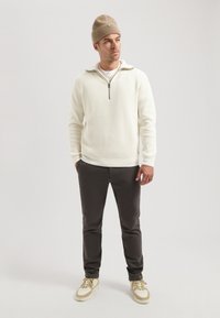 Pulover cu mânecă lungă, de culoare albă murdară, cu un guler cu jumătate de fermoar, purtat cu pantaloni închiși la culoare și adidași crem. O pălărie tricotată maro completează ținuta.