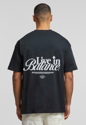 Tricou negru din bumbac, cu mâneci scurte, având pe spate un text alb mare care spune "Trăiește în echilibru", cu detalii suplimentare dedesubt.