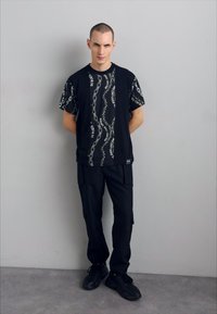 T-shirt preta com um padrão de texto branco ondulado, emparelhado com calças cargo pretas. O material parece suave. Ténis pretos, em pé contra uma parede cinza.