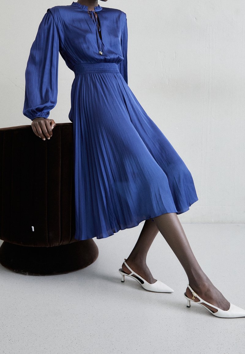 Vestido plisado azul royal con cuello alto y mangas largas, combinado con tacones blancos tipo slingback. La tela lisa contrasta con la silla texturizada.
