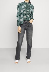Green floral-patterned blouse with ruffled neckline, paired with high-waisted gray jeans. White open-toe heeled sandals.