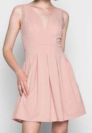 Robe rose pâle sans manches avec bretelles en dentelle, décolleté en V profond, taille cintrée et jupe plissée en ligne A portée par une personne avec une main posée sur la hanche.