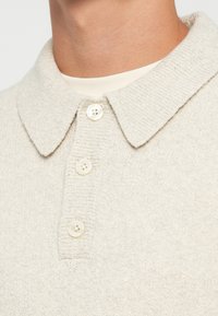 Polo de punto beige con una textura suave, que presenta un collar, cierre de tres botones blancos y detalles acanalados en el collar y la tapeta.