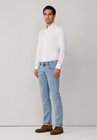 Muškarac stoji noseći bijelu košulju dugih rukava, svijetloplave traperice i smeđe ante cipele s vezicama na jednobojnoj pozadini.