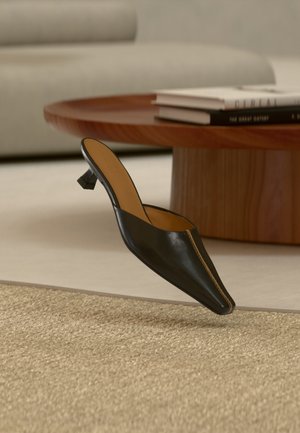 Musta nahkainen mule-mallinen kenkä, jossa on matala geometrinen korko, beige maton päällä lähellä pyöreää puista sohvapöytää, jossa on pino kirjoja.