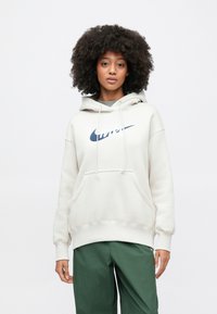 Camisola com capuz de cor creme, com bolso frontal, cordões e um proeminente logótipo da Nike em azul-marinho no peito. Tecido suave, corte descontraído.