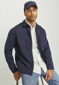 Chaqueta azul marino de botones con un bolsillo; llevada sobre una camisa blanca y azul a rayas; combinada con jeans azul claro y gorra azul marino.