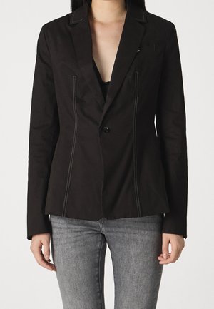 Woman wearing a fitted black blazer with white vertical stitching and a single button, paired with gray jeans against a plain background.