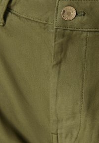 Detailní záběr na olivově zelené kalhoty, které mají hladkou tkaninu, kontrastní stehy, zlatý knoflík a detail boční kapsy.