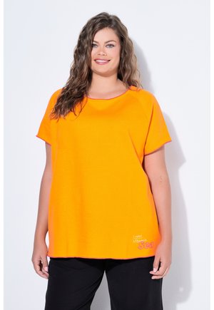 Lächelnde Frau mit welligem braunem Haar trägt ein leuchtend orangefarbenes Kurzarmoberteil mit dem Aufdruck "I need Vitamin Sea", kombiniert mit schwarzen Hosen.