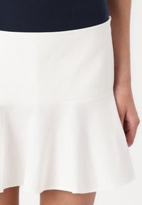 Jupe évasée blanche en tissu léger, avec une texture lisse et une taille ajustée. L'ourlet finit par une courbe subtile.