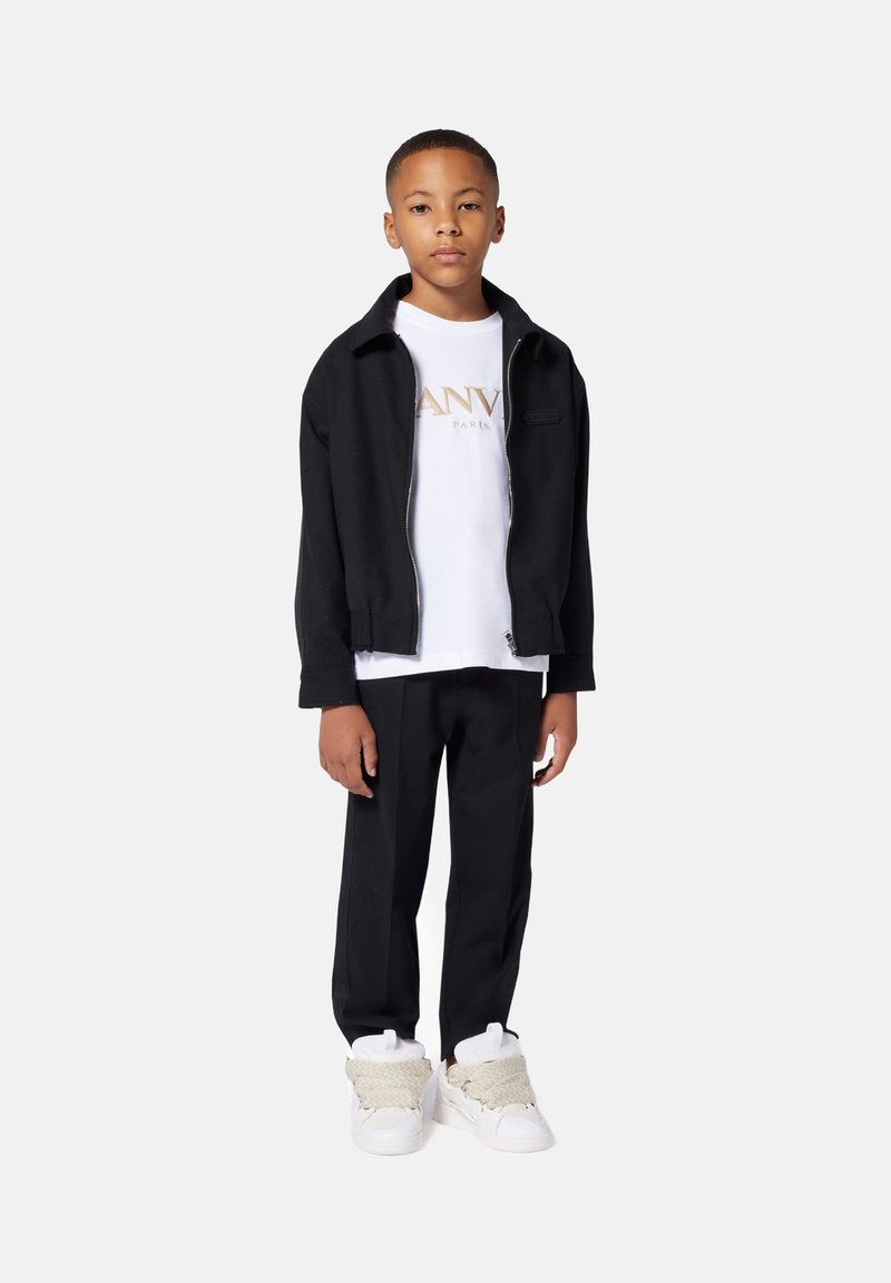 Zwarte zip-up jack over een witte T-shirt met gouden letters, gecombineerd met zwarte broeken en witte sneakers met gestructureerde accenten.