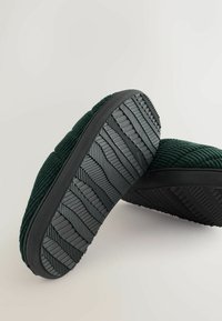 Pantofole in corduroy verde con superficie testurizzata e suole in gomma nera che presentano un motivo a zig zag per migliorare la presa e la stabilità.