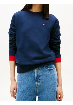 Kvinde iført marineblå sweatshirt over en synlig rød langærmet skjorte, kombineret med lyseblå højttaljede jeans.