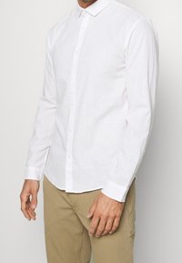 Camicia bianca a maniche lunghe realizzata in un materiale simile al lino, dotata di chiusura con bottoni, colletto e polsini arricciati. Texture liscia e leggera.