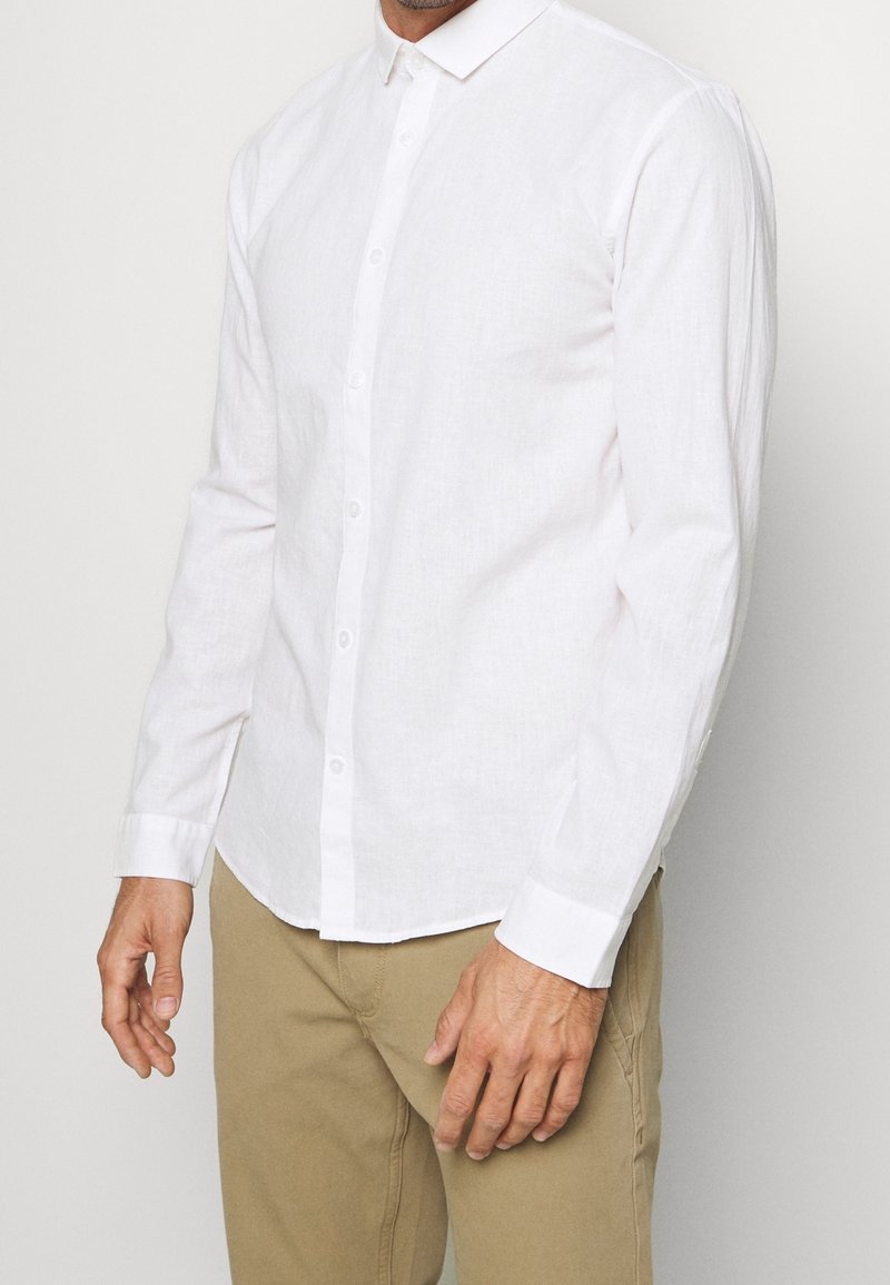 Camicia bianca a maniche lunghe realizzata in un materiale simile al lino, dotata di chiusura con bottoni, colletto e polsini arricciati. Texture liscia e leggera.