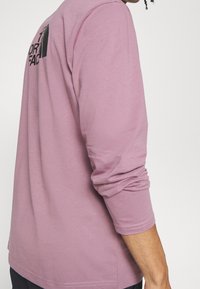 Tričko s dlouhými rukávy v jemné růžové barvě z měkké bavlny, s kulatým výstřihem a černým grafickým logem na zadní straně. Hladká textura s prošitými lemy.