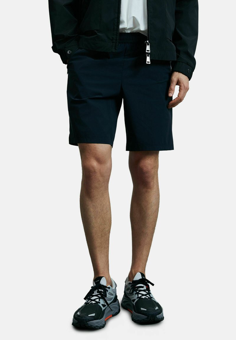Un short bleu marine à coupe décontractée, avec une taille élastique et des poches latérales, associé à des chaussures de sport noires et grises avec des accents orange.