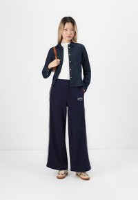 Jeansjacka, vit skjorta, marinblå vida sweatpants med röda ränder och mönstrade sneakers. Utrustad med en axelväska och avslappnad passform.