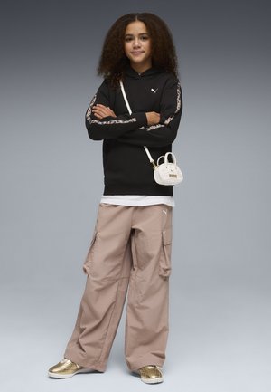 Junge Person mit lockigem Haar, die einen schwarzen Hoodie, beige Cargo-Hosen, goldene Sneakers und eine kleine weiße Umhängetasche trägt, stehend mit verschränkten Armen.