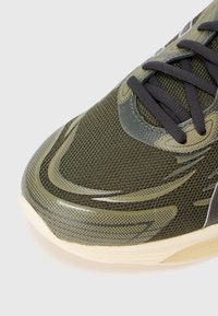 Olivza zaļa sporta kedu ar sieta materiālu, teksturētu virsmu, gludām akcentiem, melnām šņorēm un baltu gumijas zoli ar rakstu detaļām.